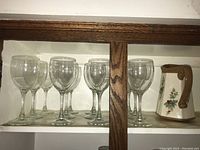 Set of twelve clear wine glasses displayed on left side of cabinet shelf with one floral decorated ceramic jug with brown handle on right.