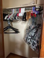 A closet rod holding assorted adult plastic hangers in white, black, blue, and red, wooden hangers, and colorful small-sized kids hangers. A large plastic bag on the floor contains additional hangers.
