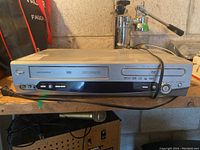 Front view of Magnasonic DVD825 combination VCR and DVD player showing VHS tape slot, DVD tray, control buttons and cable coiled on top.