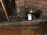 Photo of oil lamp, metal magazine rack, brass decorative cow bell, small candle holder, and black metal lantern on a hearth in front of brick.