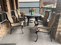 Six metal framed patio chairs arranged on porch around a round glass-top table with a flower pot centerpiece.