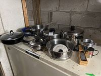 Various stainless steel pots and pans with lids, a Bialetti casserole pan with lid, stainless steel cups, a handheld grater, arranged on a surface.