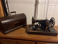 Black vintage Singer electric sewing machine with floral decals displayed on wooden surface next to its brown removable carrying case with metal handle and Singer logo.