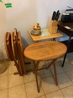 Photo showing four oval wooden TV tables with foldable legs hanging on a wooden stand, a single wooden folding tray, and a wooden knife block with kitchen knives on a small table.