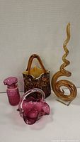 Amber spiral glass sculpture, cranberry glass bag, cranberry square basket, and bud vase arranged; highlights shapes and colors