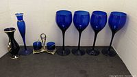 Four large cobalt blue wine glasses, one slender cobalt blue glass bud vase, one silver condiment holder with two blue glass containers, and one black and gold ceramic bud vase arranged on a black surface against a white background.