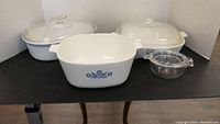 Four casserole dishes on display: three white Corning Ware with blue cornflower pattern and lids, one small clear Pyrex glass dish with lid