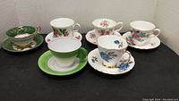 Set of six various bone china cups and saucers displayed on black surface against white background.