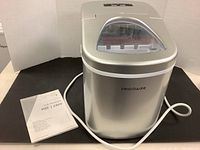 Silver Frigidaire ice maker with user manual and power cord beside it on a black surface.