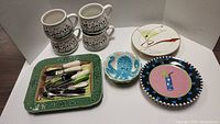 Four large cow pattern soup mugs stacked, 4 decorated martini side plates visible, green asparagus serving plate with assorted flatware