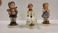 Front view of 3 Hummel figurines, showing boy with fruit, girl with geese, and girl with flowers.