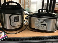 View of Instant Pot Duo and DeLonghi crock pot side by side on a shelf with cords visible and user manual partially shown.