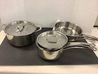 Five cookware items including large stock pot with lid, two saucepans with lids, and two frying pans shown on a table.