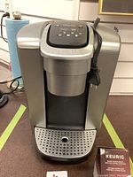 Front view of silver Keurig coffee maker with power cord wrapped around the side, drip tray at bottom, and brand name on front.