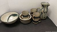 Grouped view of entire set showing 6 saucers stacked, 5 cups stacked and standing, ceramic pitcher with blue vine design, and bowl with brush strokes.
