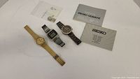 Three men's watches arranged with manuals and a small bag of extra parts, showing gold Seiko, black/silver digital Seiko, and a Michael Kors watch with a bling bezel.