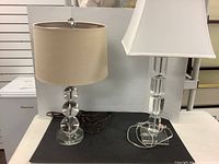 Two glass table lamps placed side by side on a black surface with visible power cords and shade details.