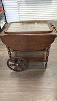 Front view of vintage wooden tea trolly with glass tray on top, drawer underneath, and fold-down side leaves.