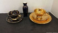 Overall photo showing all items together: two teacup and saucer sets (one cobalt blue and gold, one gold-colored), a gold-colored plate, and a dark blue and gold vase.