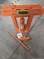 Full view of the 12 ton hydraulic pipe bender with orange frame, hydraulic ram, bending die, and handle on the floor.