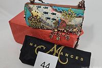 Overall view of the Mary Frances handbag resting on a red cloth, showing the full shape and the carrying strap along with the brand dust bag and lot tag.