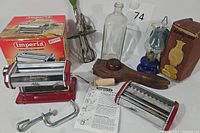 Full view of the lot showing Imperia pasta machine, vintage bottle, wooden citrus squeezer, egg beater, and mini oil lamp in box.