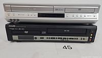 Two VHS/DVD combo players stacked, Toshiba on top (silver finish) and Philips below (black finish). Visible control buttons and ports.