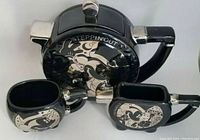 Front view of teapot, creamer, and teacup all featuring black ceramic with silver Mickey Mouse Steppin Out graphics and silver highlights on handles and spouts.