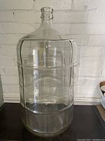 Clear 6 gallon glass wine carboy shown against a white brick wall, sitting on a black surface. The carboy is transparent with ribbed vertical sides and a narrow neck.