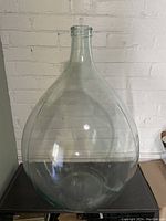 Clear glass wine carboy standing against a white brick wall, showing the full shape and size of the item.