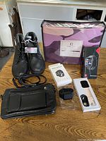 Photo showing all items in lot on wooden table including black boots, bed sheet package, two smart watches, and black shoulder bag.