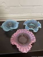Three glass hobnail pattern candy dishes, two blue and one pink, arranged on a black surface against a white painted brick wall background.