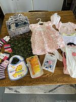 Lot of baby clothes and accessories on table, including pink dress, onesies, bib, socks, feeding utensils.
