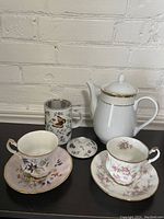 Two Paragon tea cup and saucer sets, one Teaopia tea cup with steeping strainer, and one white ceramic tea pot shown together on a black surface with a white brick background.
