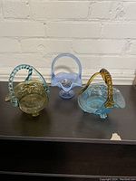 Three glass baskets displayed side by side showing different colors and designs, two with reversed color handles, one opalescent blue basket.