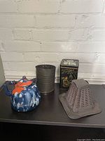 Lot of four vintage kitchenware items displayed against a white brick wall: ceramic teapot, metal sifter, decorated tea tin, vintage toaster rack