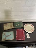 Photo showing three vintage metal advertising tins and two metal beer ashtrays with a lighter on right side. Tins and ashtrays display advertising graphics and exhibit signs of wear and age.
