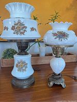 Two vintage milk glass lamps with metal bases, floral patterns, original cords, photographed on the table with plants in background.