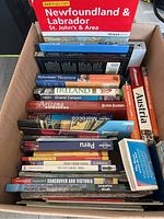 Photo of a box filled with various travel books and atlases, titles visible including Newfoundland & Labrador, Ireland, Grand Canyon, Arizona, Morocco, Peru, Austria.