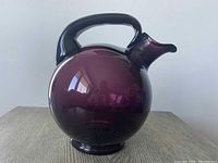 Front view of the rounded amethyst purple glass pitcher with an arched handle and spout on a wooden table, showing overall form and color.