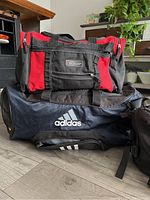 View of two duffle bags stacked: Adidas navy blue duffle bag with white logo and Dunlop red and black duffle bag.