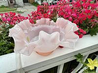 Side angled photo showing the ruffled, pink glass bowl placed on a white railing outdoors with pink flowers in the background.