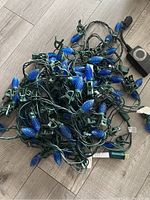 Blue textured baseball bat-shaped LED Christmas lights tangled with green wiring, clips attached to wiring, lying on a light wood floor.