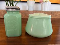 Side by side view of a jadeite glass shaker and a lidded jar on a wooden surface in front of a shelf.
