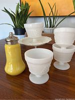 Yellow shaker with perforated metal top placed alongside three custard cups and a yellow metal shaker stand on a wooden surface, with plants in the background.