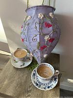 Photo of two vintage tea cups with saucers, each holding a beeswax candle, shown next to a purple beaded silk hanging light swag decorated with embroidered flowers and butterflies.