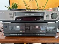 Front view of Toshiba VHS player on top and Technics dual cassette deck below on wooden surface with plants in background