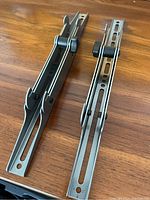 Two metal TV mount hooks laid out on a wood surface showing the full length and slots for mounting adjustments.