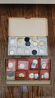 Top-down view of two boxes containing various watch faces and metal watch cases. Several watch faces are stored in a plastic tray slots, and cases are arranged in red cardboard compartments with some faces sealed in plastic.