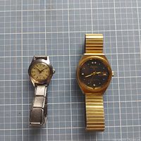 Two vintage wristwatches laid side by side on a grid background. One is a silver stainless steel watch with silver dial, the other is a gold-tone Longines 23 with black dial and an expansion band.